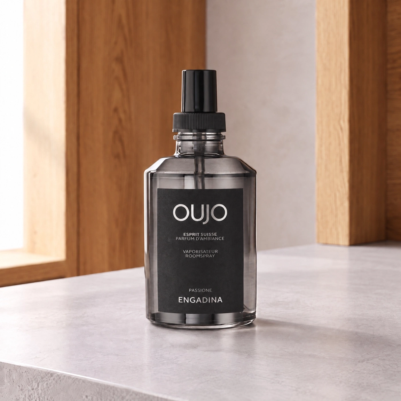 A bottle of "OUJO" room spray with a black label and silver text. The background is blurred, showing wood. A simple, elegant product photo.