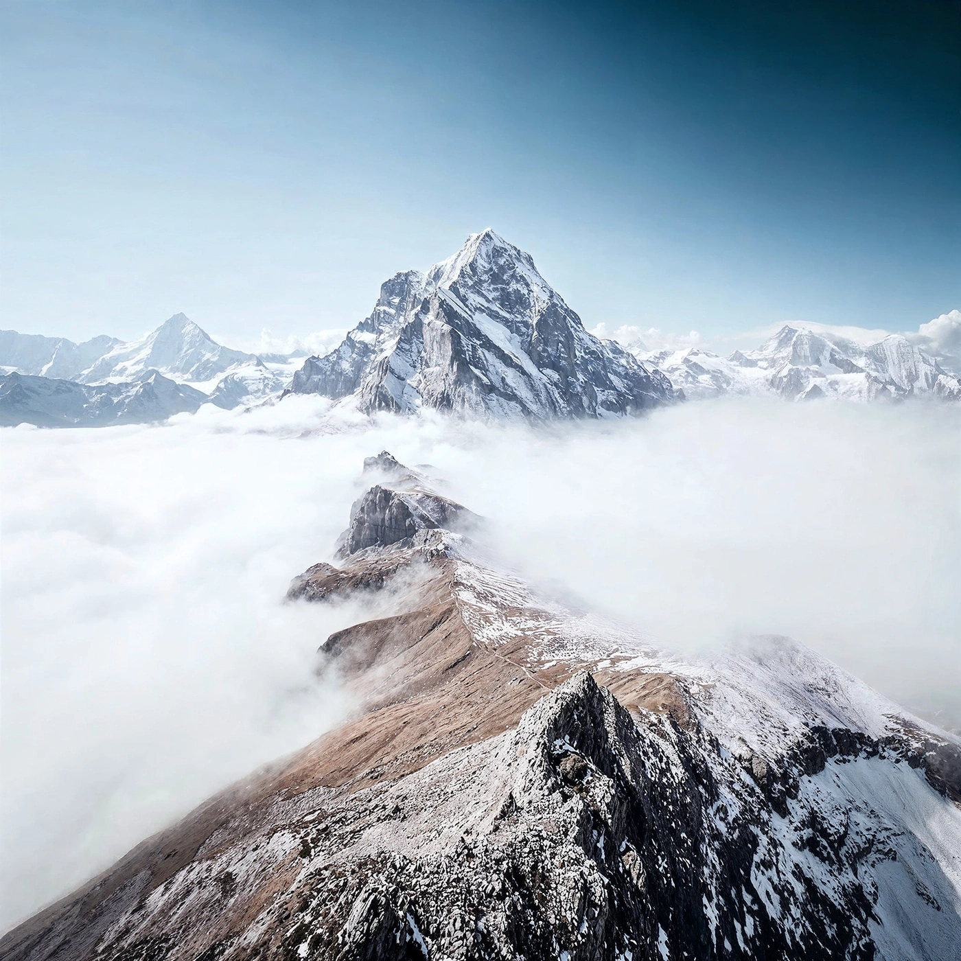 Snow-covered mountain peaks rise above dense clouds under a clear sky.