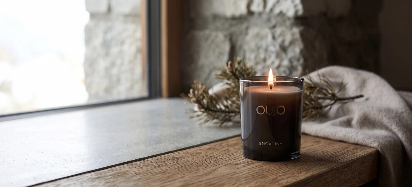 Burning candle labeled 'Engadina' on a windowsill next to a cozy blanket.