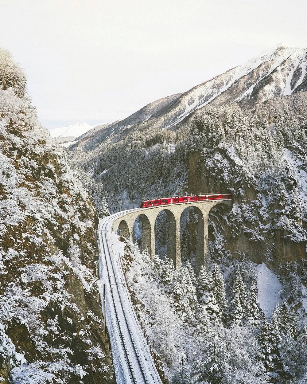 Red train crossing a bridge in a snowy mountain landscape.