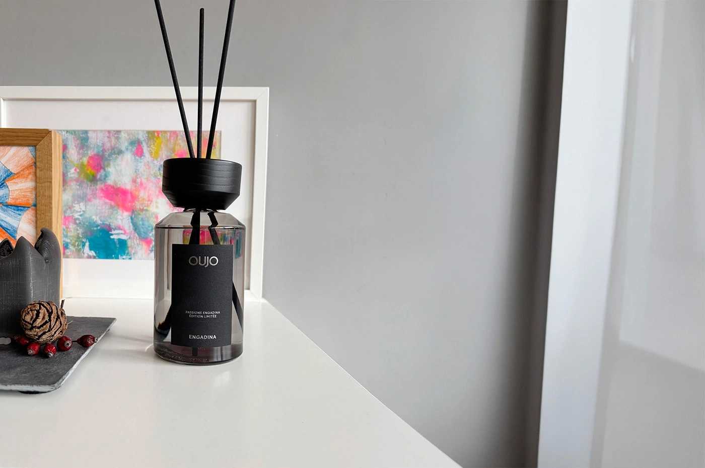 A diffuser with a black label and three reeds stands on a white table.