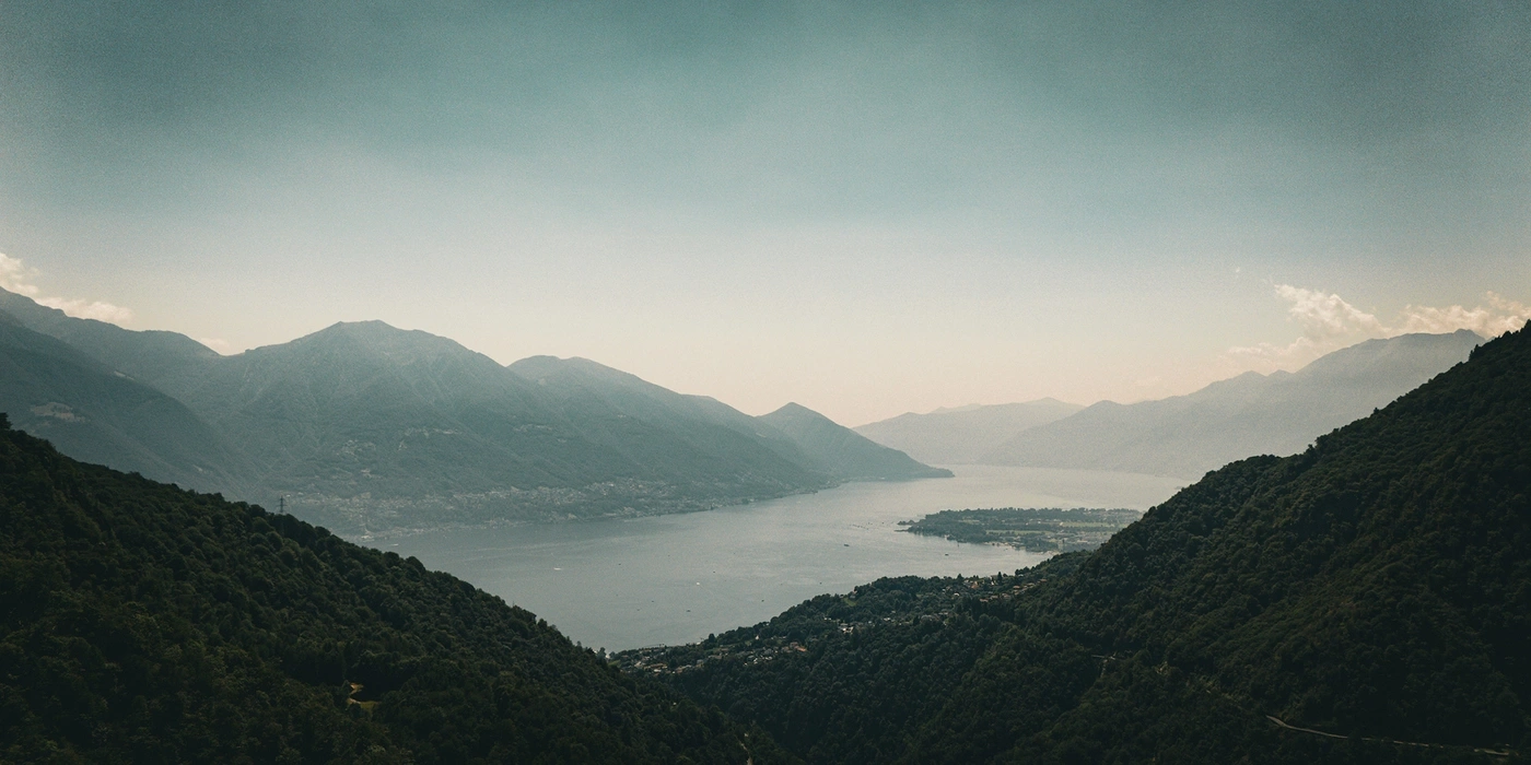 Mountain landscape with a lake, forested slopes, and a hazy sky.